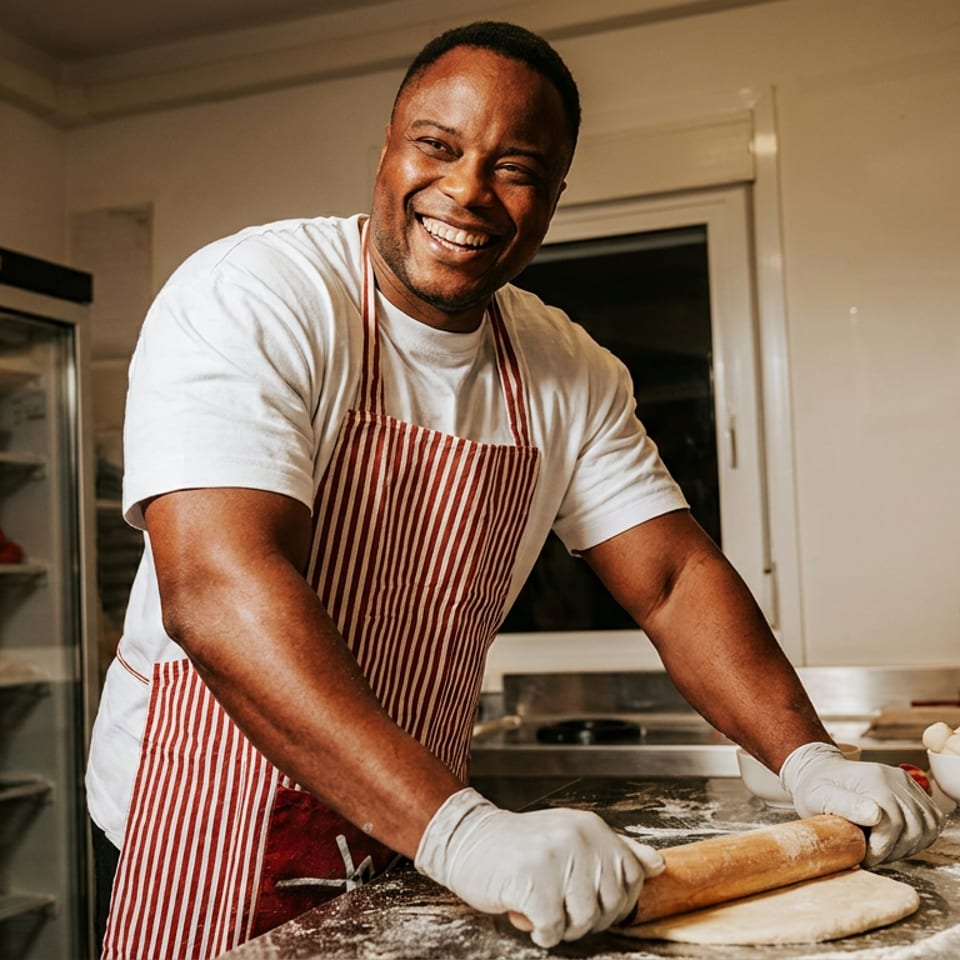 Smiling chef in a striped apron and gloves rolling dough on a floured counter in a commercial kitchen.
